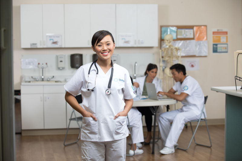 Nursing student wearing white scrubs and a stethoscope. Three other students talking together in background.